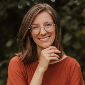 Alyssa Schwarz, Frau mit Brille und dunklen Haaren vor hellem Naturhintergrund lächelnd