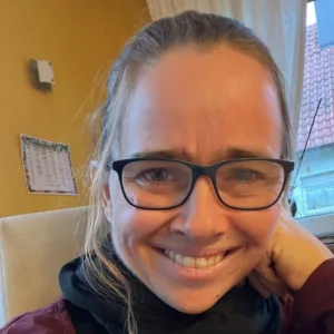 Judith Oesterle, Sprecherin mit Brille und langen Haaren in dunklem Shirt vor gelbem Hintergrund