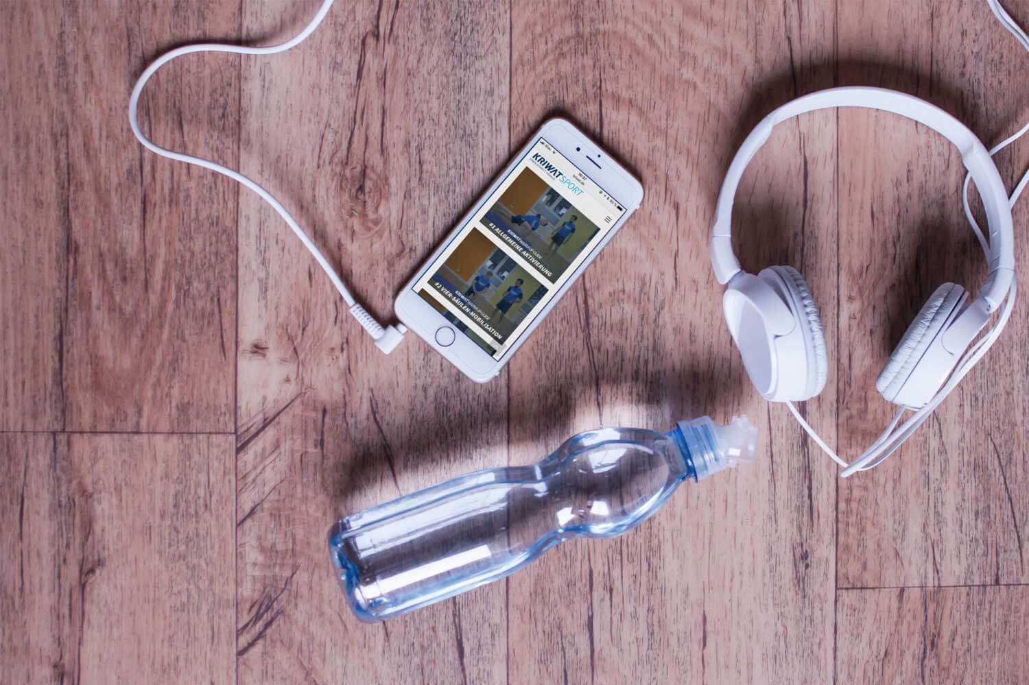 Kriwat Warm-Up-Guide – Videokurs zum Aufwärmen von Kriwat GmbH 1 Smartphone mit Kriwat-Kurs, Kopfhörer und Wasserflasche auf Holztisch