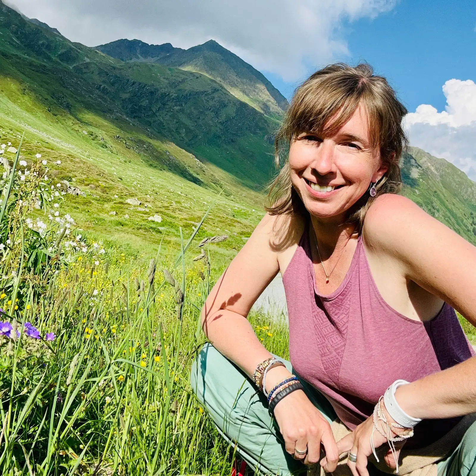 Frau in rosa Top lächelt in grünem Bergtal mit weißen Blüten im Gras