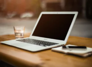 Weißes MacBook auf Holztisch mit Notizbuch, Stift und Wasserglas, unscharfer Büro-Hintergrund