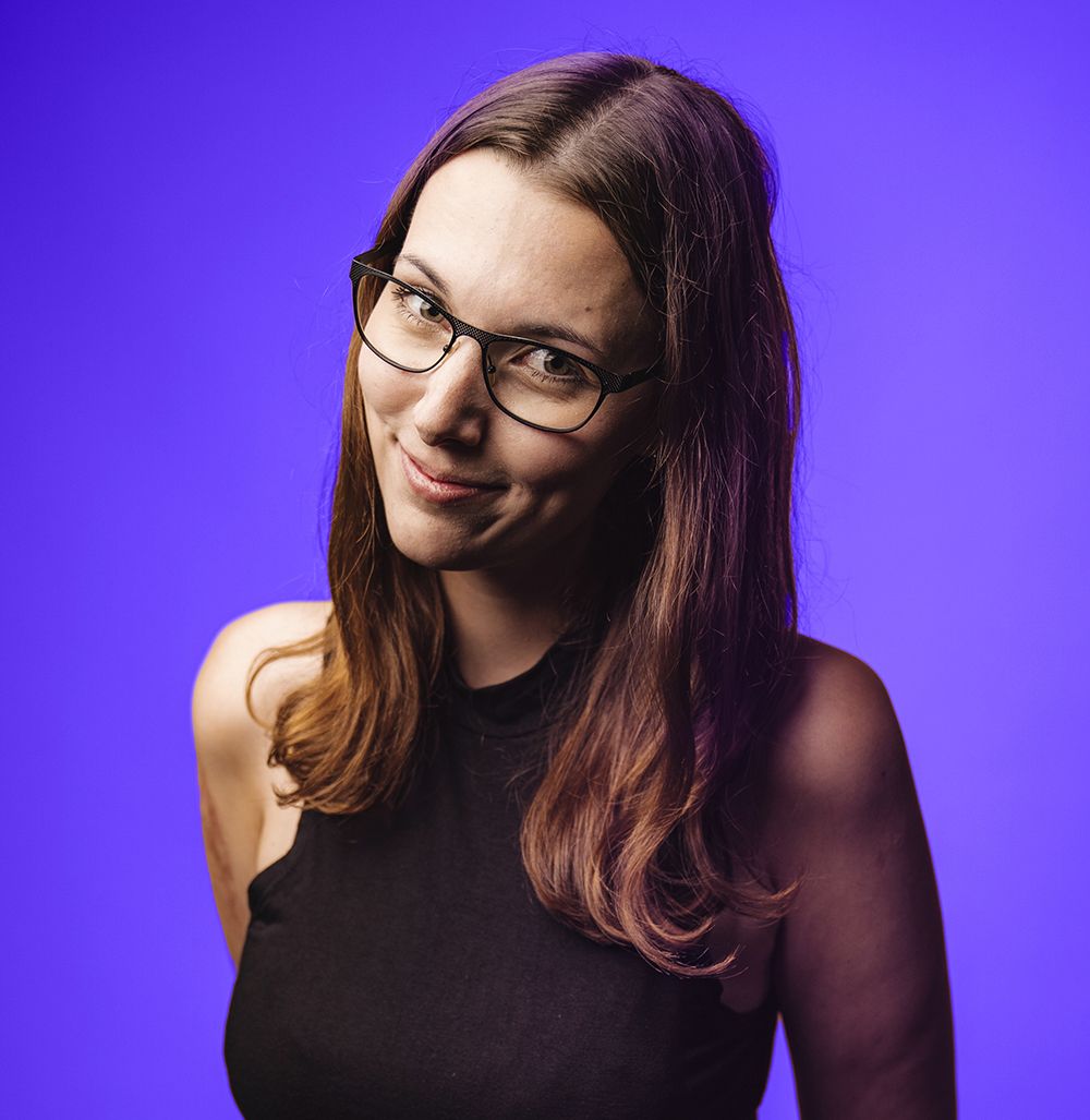 Porträtfoto einer Frau mit Brille und dunklen Haaren vor lila Hintergrund