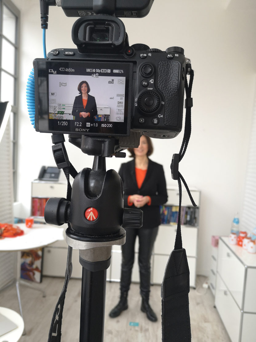 Sony-Kamera auf Stativ mit Frau in schwarzem Blazer und orangerrotem Schal im Hintergrund während Video-Aufnahme