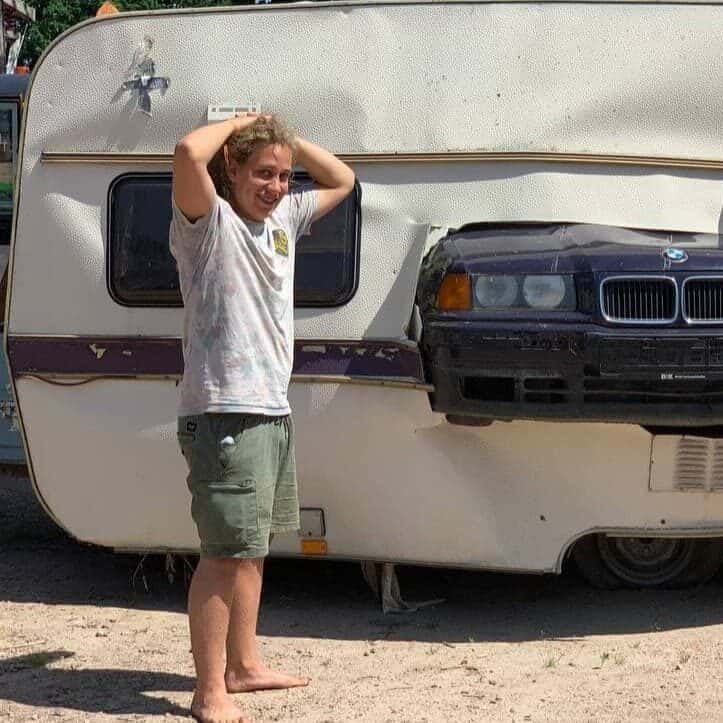 Junger Mann in hellem T-Shirt vor Wohnwagen, hält sich Hand an Kopf