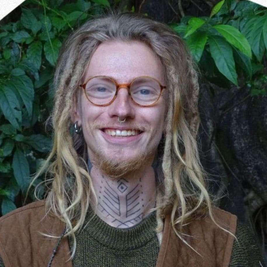 Porträt eines lächelnden Mannes mit langen Haaren, Brille und Halskette vor grünem Hintergrund
