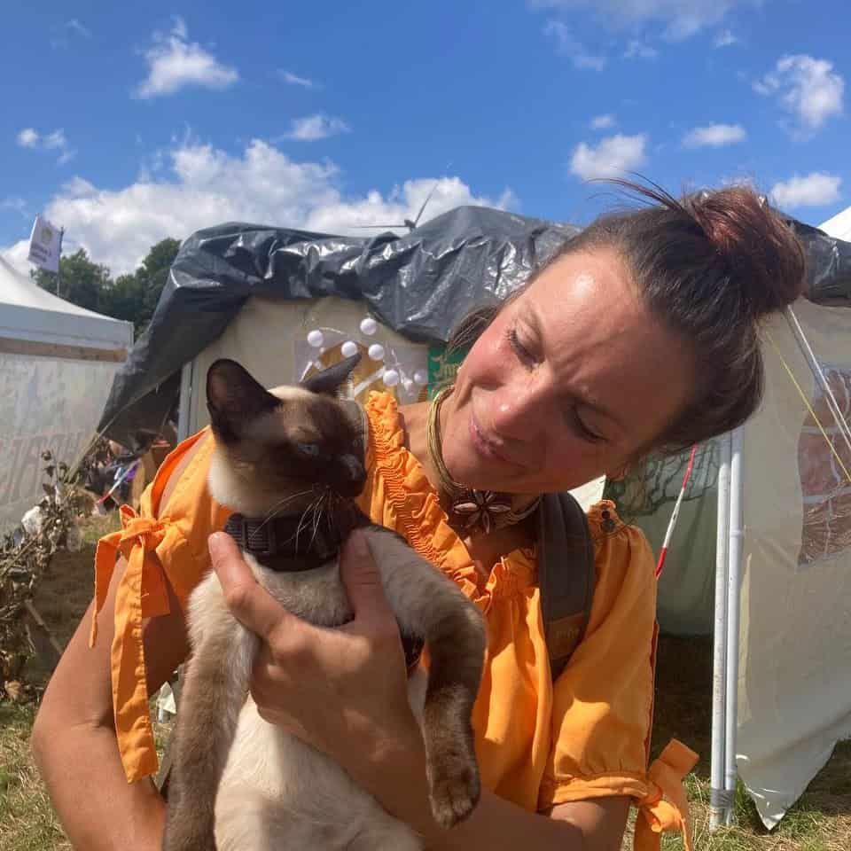 Frau in orangefarbener Jacke hält Katze in den Armen vor hellem Himmel