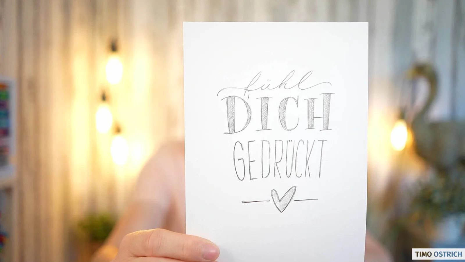 Skizze 'Fühl dich gedrückt' mit Herzchen auf Papier vor verschwommenem Hintergrund mit Lichtern