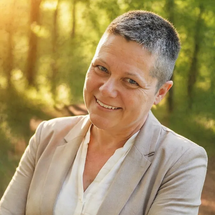 Karin Zeilinger lächelndes Porträt in Beige-Blazer vor grünem Waldwald-Hintergrund