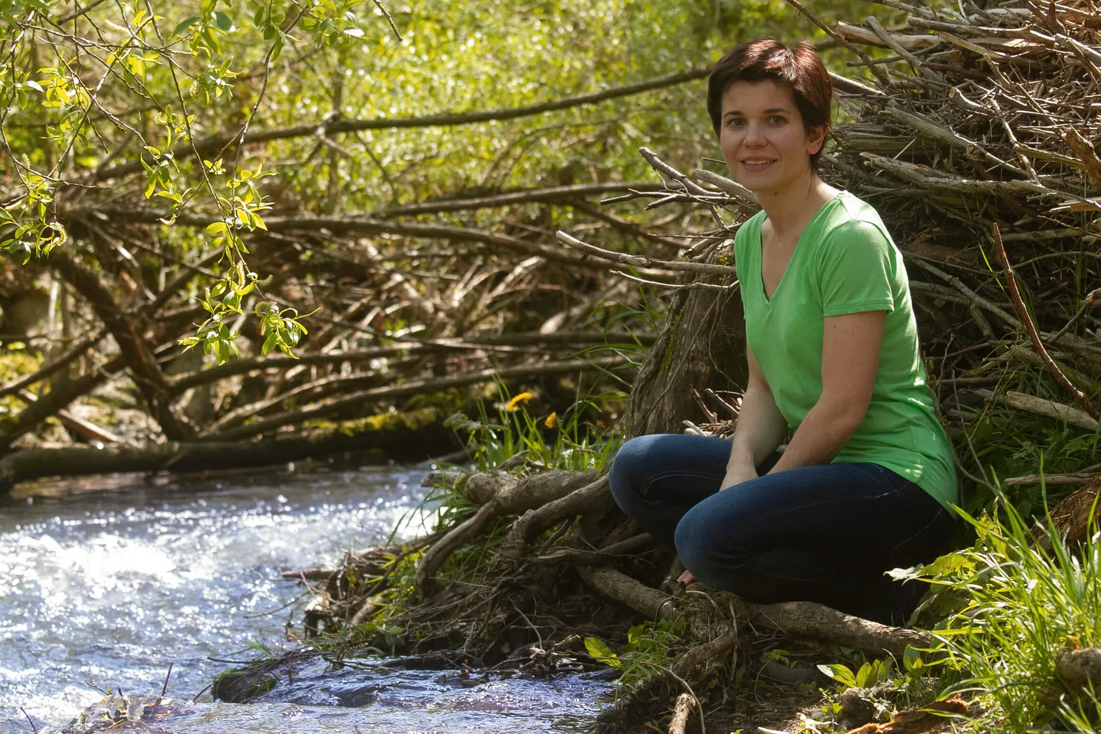 Natalie Klug in grünem Shirt hockt am Bachufer zwischen Ästen und Weidenbäumen