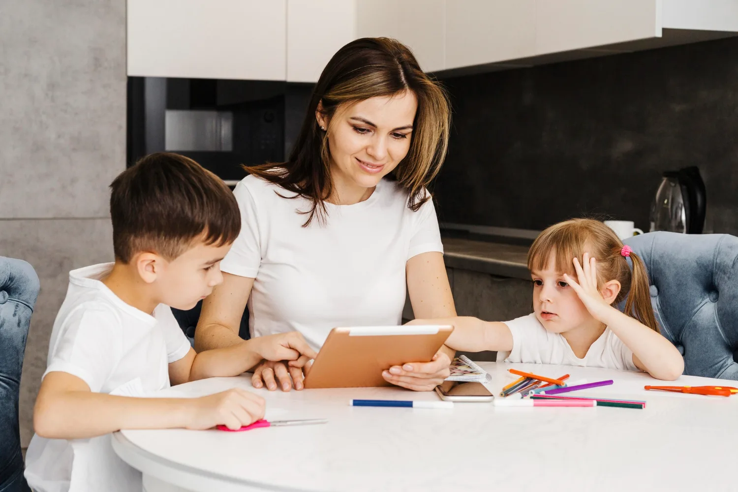 Mutter mit zwei Kindern am Tisch mit Tablet und Buntstiften, moderne Küche im Hintergrund