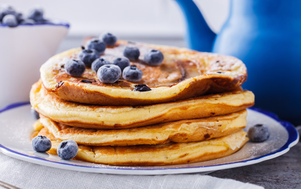Low Carb – Abnehmkurs mit Ernährungsplan von OnVersum GmbH 4 Stapel von Pfannkuchen mit Blaubeeren auf weißem Teller mit blauem Krug im Hintergrund