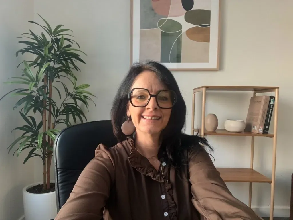 Beatrix sitzt am Schreibtisch in ihrem Büro mit Topfpflanze, Bild und Regal im Hintergrund