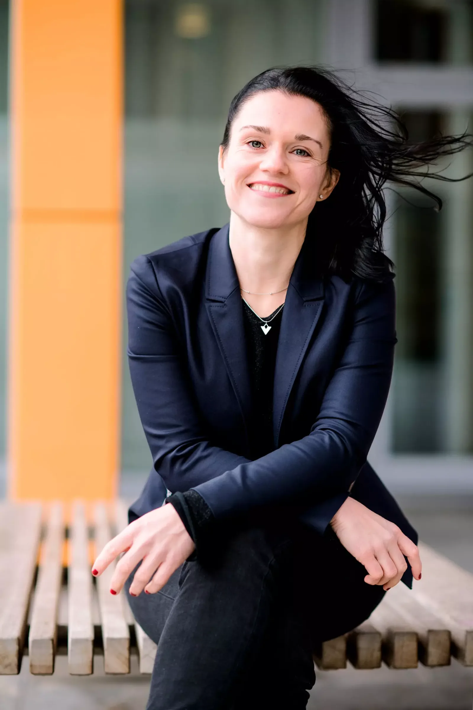 Frau mit dunklen Haaren in dunklem Blazer sitzt vor orangem Balken, lächelnd