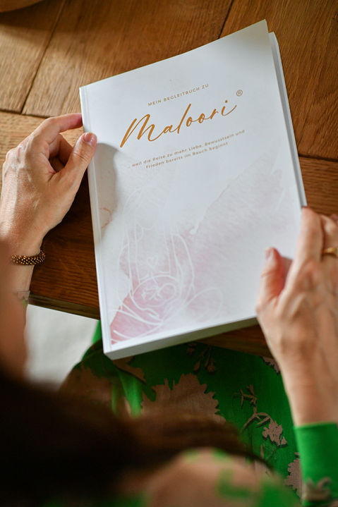 Frau hält Maloori Begleitbuch mit goldenem Schriftzug und Rosa-Illustration