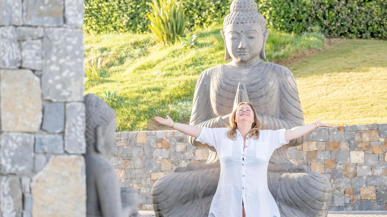 Master of Communication – Kommunikations-Coaching 3 Frau in weißem Hemd mit ausgebreiteten Armen vor Buddha-Statue in Garten, entspannte Haltung