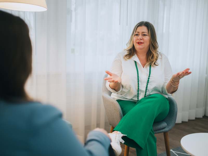 Master of Communication – Kommunikations-Coaching 4 Frau in Coachinggespräch, sitzt mit grüner Hose und weißer Bluse in moderner Raum, gestikulierend