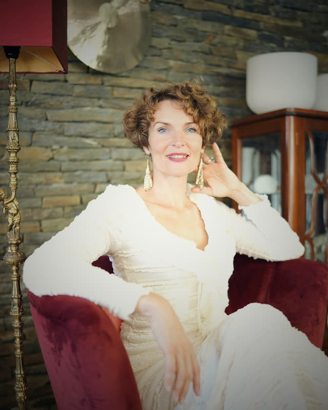 Stefanie Bruns sitzt in weißem Kleid auf rotem Stuhl in elegantem Innenraum mit Steinwand