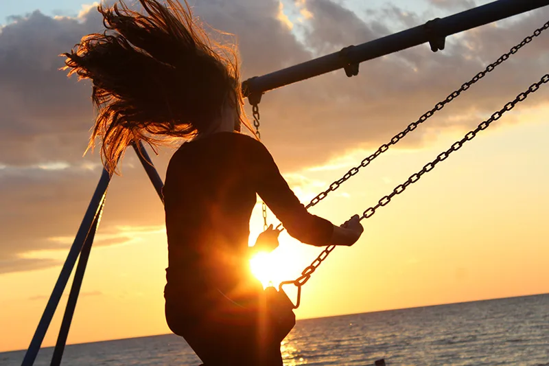 Silhouette einer Frau auf Schaukel vor Sonnenuntergang über dem Meer, goldenes Licht