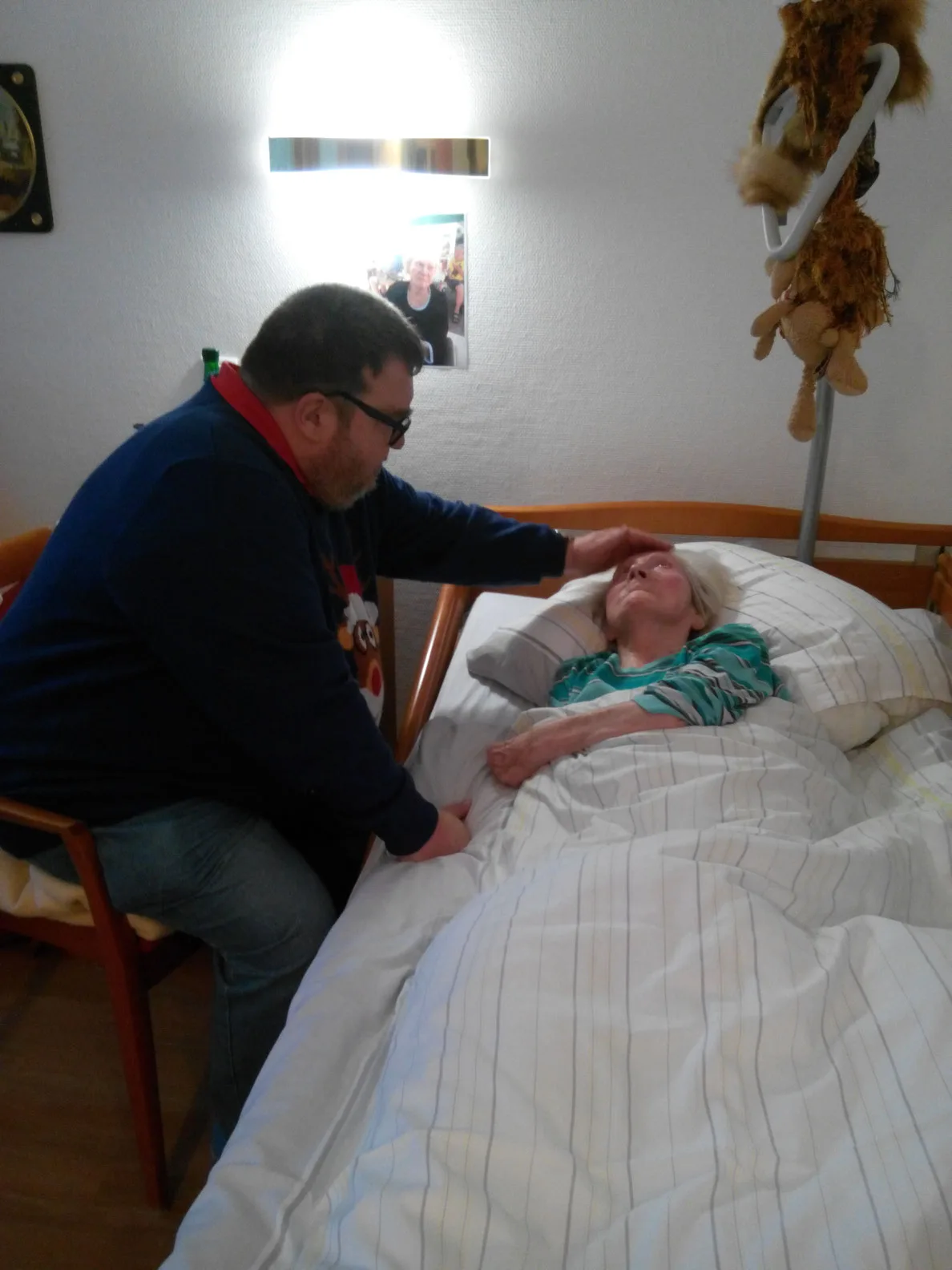 Besuchssituation: Mann sitzt neben verstorbener Person auf dem Bett, berührt sie