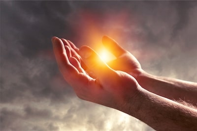 Zwei Hände halten glühendes Licht gegen bewölkten Himmel