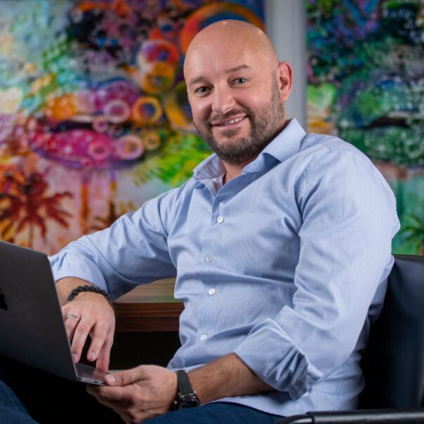 Marcel Schlee sitzt mit Laptop vor buntem abstraktem Kunsthintergrund, trägt hellblaues Hemd