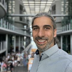 Daniel Dirks Portrait im Freien vor verschwommenem Architektur-Hintergrund, trägt helles Hemd