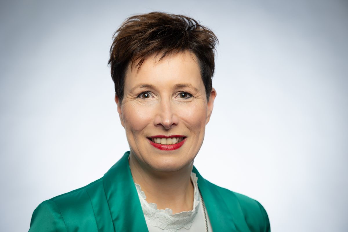 Porträt Katina Hacker mit grünem Blazer, kurzen dunklen Haaren und rotem Lippenstift vor hellem Hintergrund
