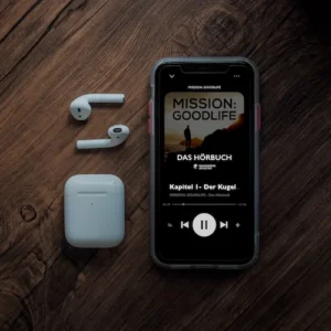 Mission: Goodlife Hörbuch-Cover auf Smartphone-Display mit AirPods und Charging Case auf Holzuntergrund