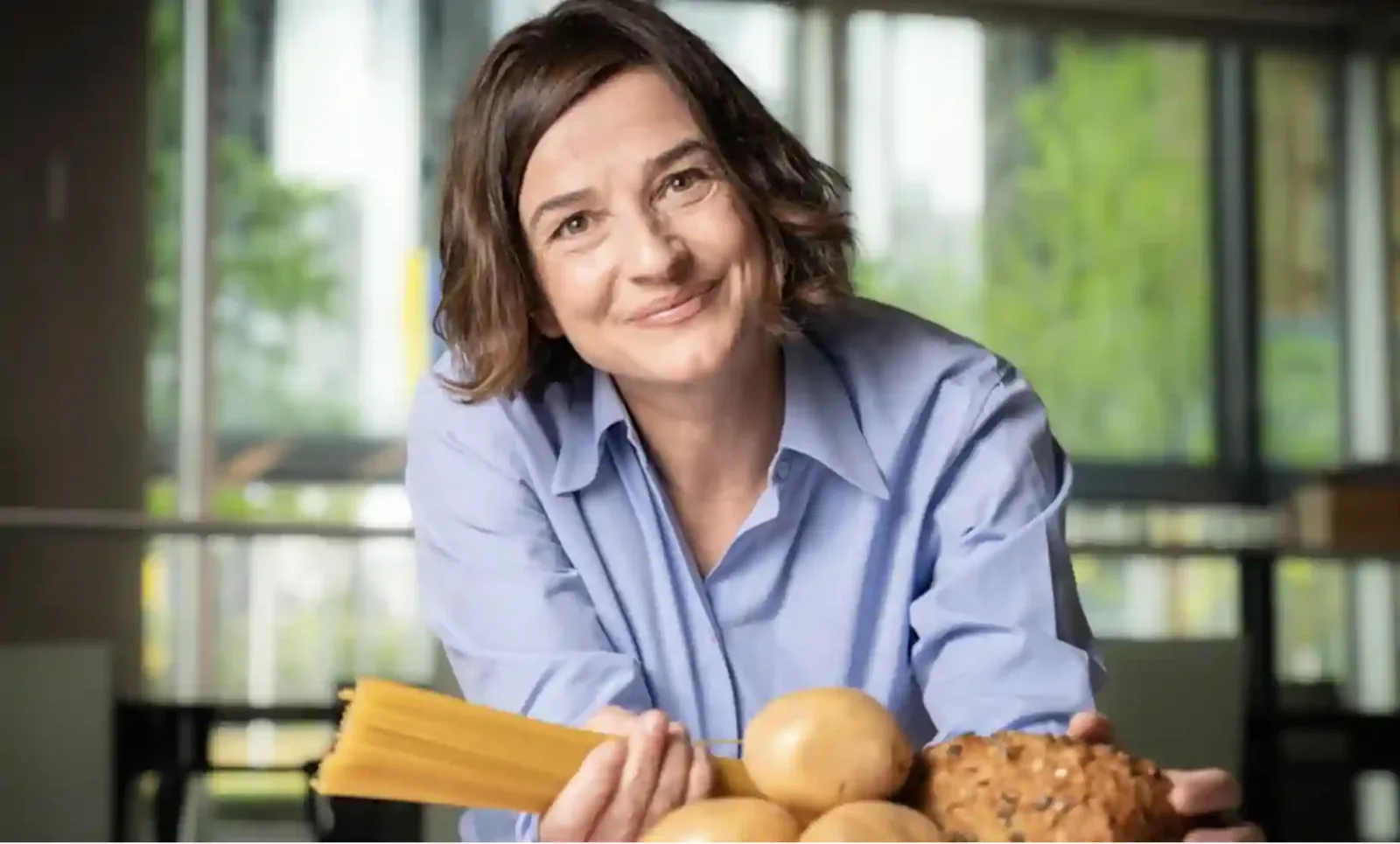 Frau in blauem Hemd hält Pasta, Kartoffeln und Brot, lächelt in die Kamera