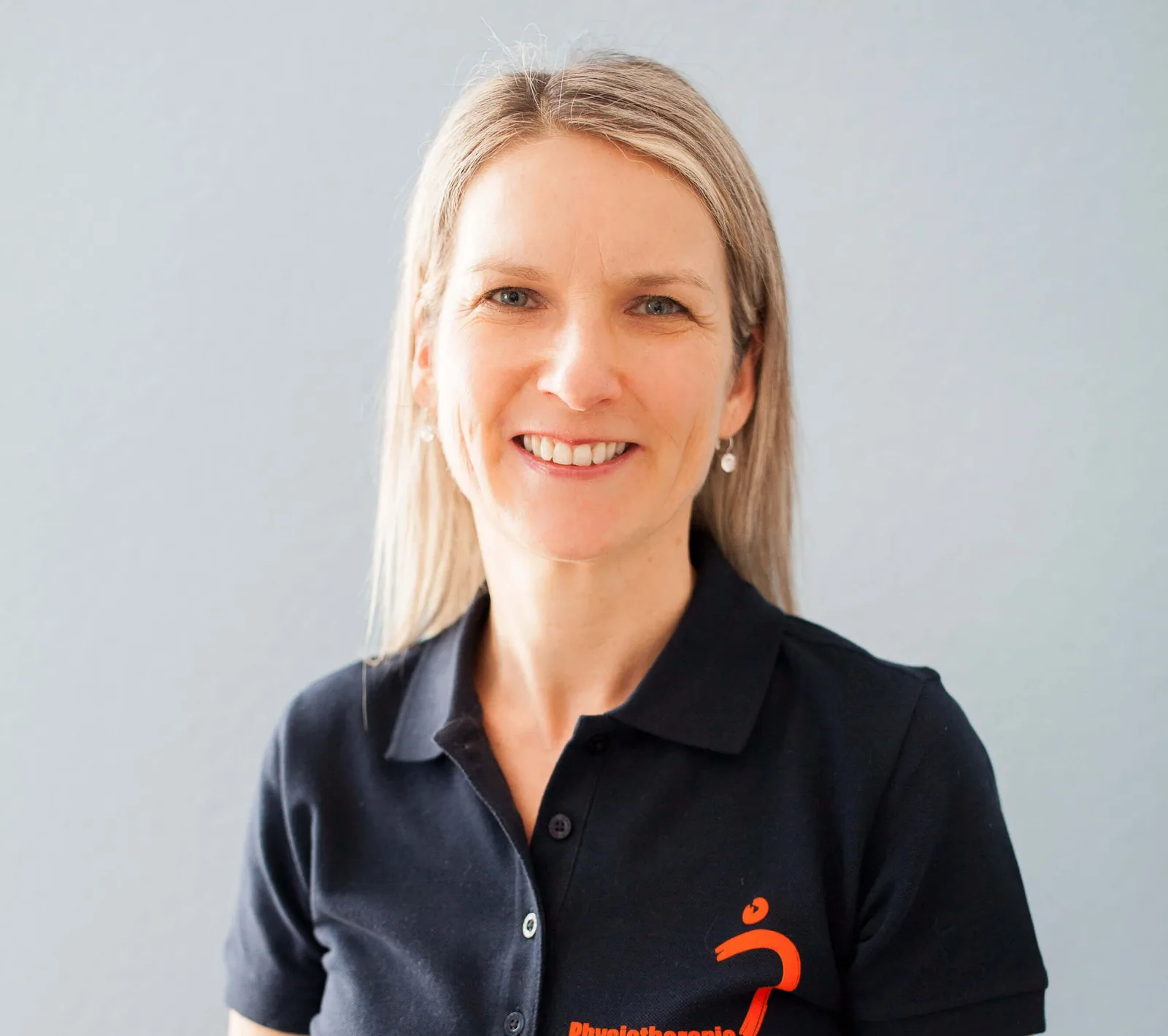 Porträtfoto von Kristin Schirbock, blonde Frau mit dunklem Poloshirt mit orange Logo, vor neutralem Hintergrund
