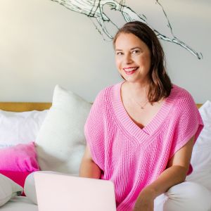 Porträt Simone Stalberg, Frau in rosa Strickpullover mit Laptop auf Bett sitzend