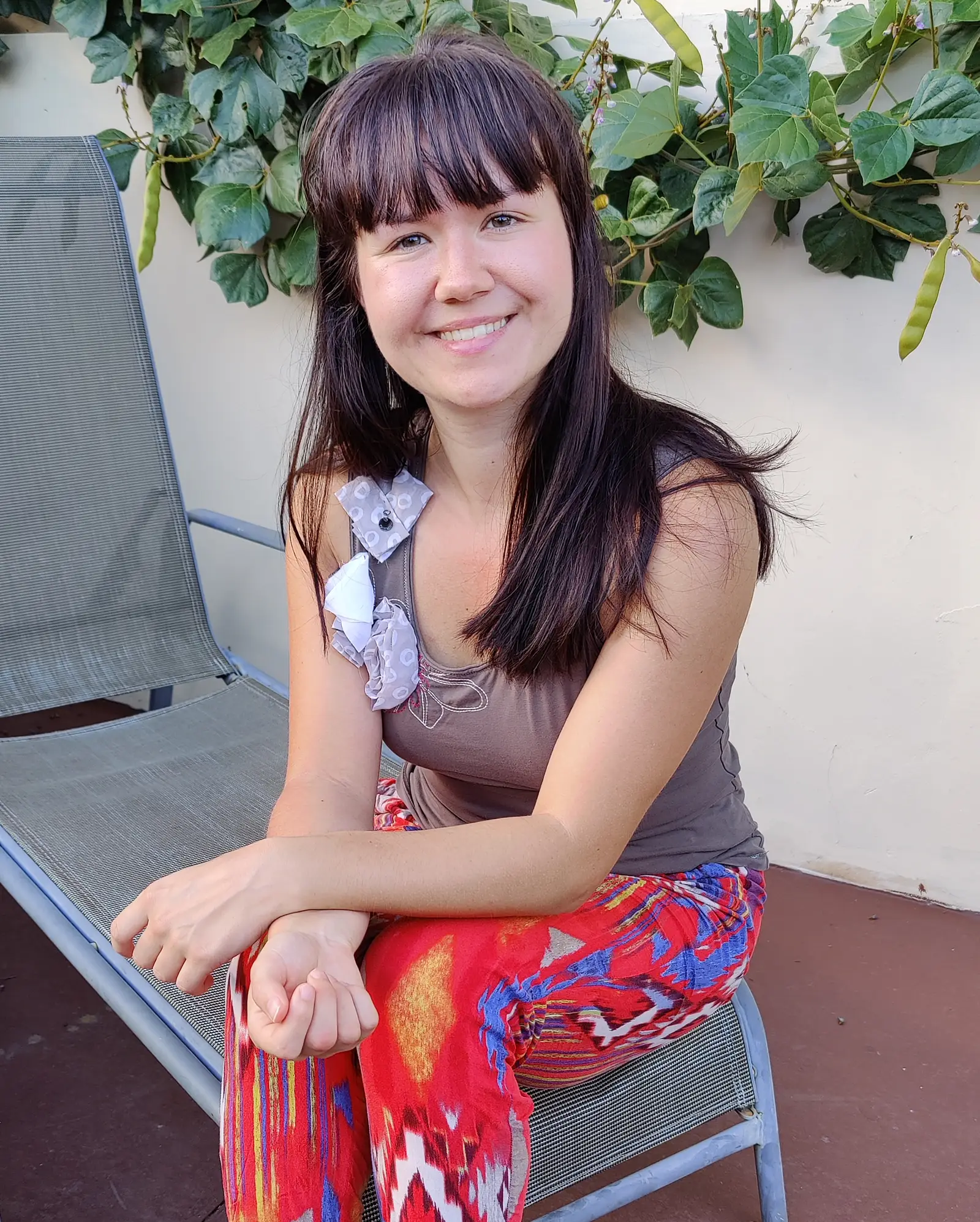Porträt einer lächelnden jungen Frau mit dunklen Haaren und Pony an Pflanze, trägt bunte rot-blaue Hose und graubraunes Top
