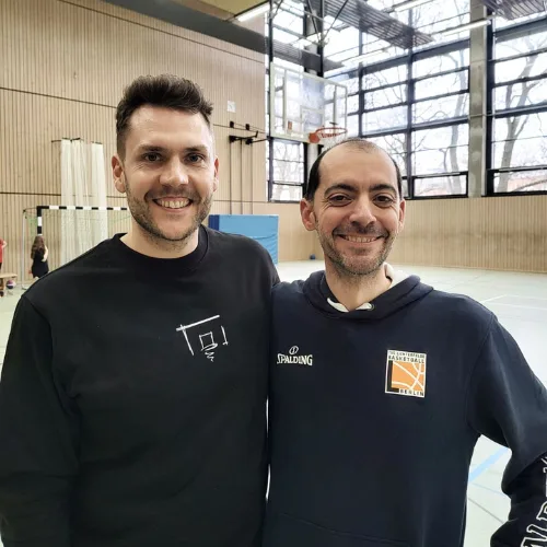 Zwei Athletik-Trainer lächelnd in Sporthalle, einer trägt schwarzes Shirt mit Trainingsprogramm-Logo