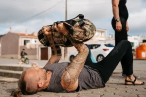 Mann mit Tattoos führt Sandsack-Wurf im Liegen durch, beobachtet von Person daneben