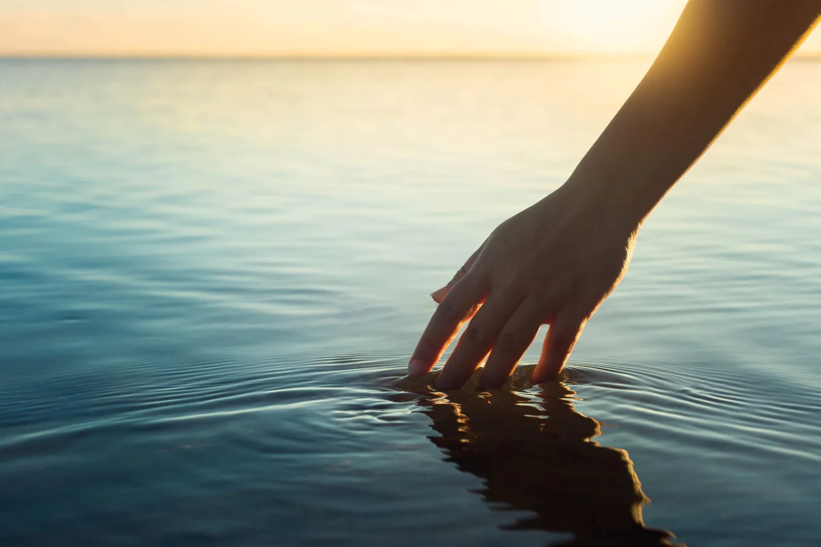 Hand berührt ruhiges Meerwasser bei Sonnenuntergang mit goldenen Lichtstrahlen
