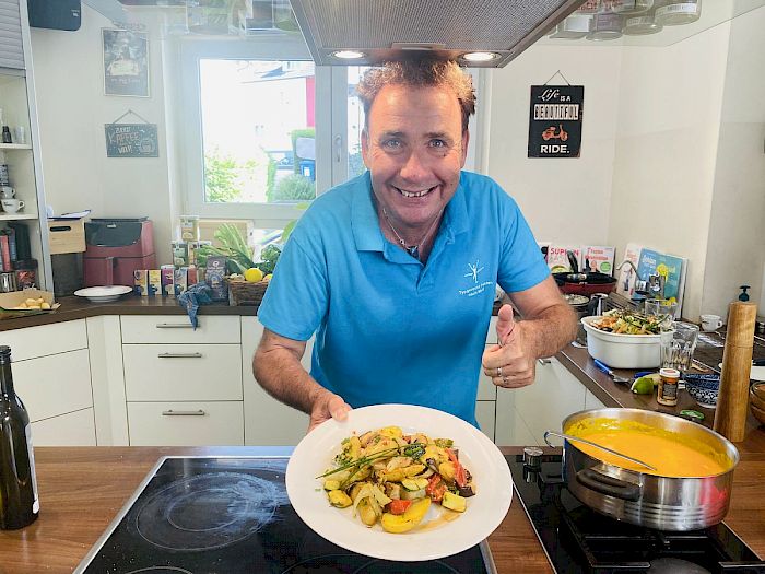 Neustart Ernährung – Basisch-Kochen-Kurs von Ralf Moll 15 Ralf Moll in blauem Poloshirt in der Küche präsentiert ein Gemüsegericht, lächelt in die Kamera