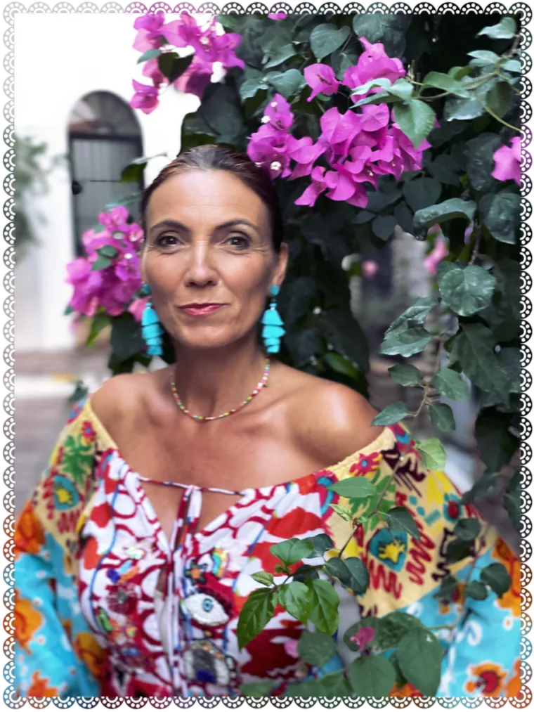 Carmen Duffner, Autorin, porträtiert vor pinken Bougainvillea-Blüten in farbenfroher Kleidung mit türkisen Ohrhängern