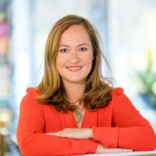 Porträt Anna Nguyen, Frau mit rotem Blazer lächelnd vor blauem unscharfem Hintergrund