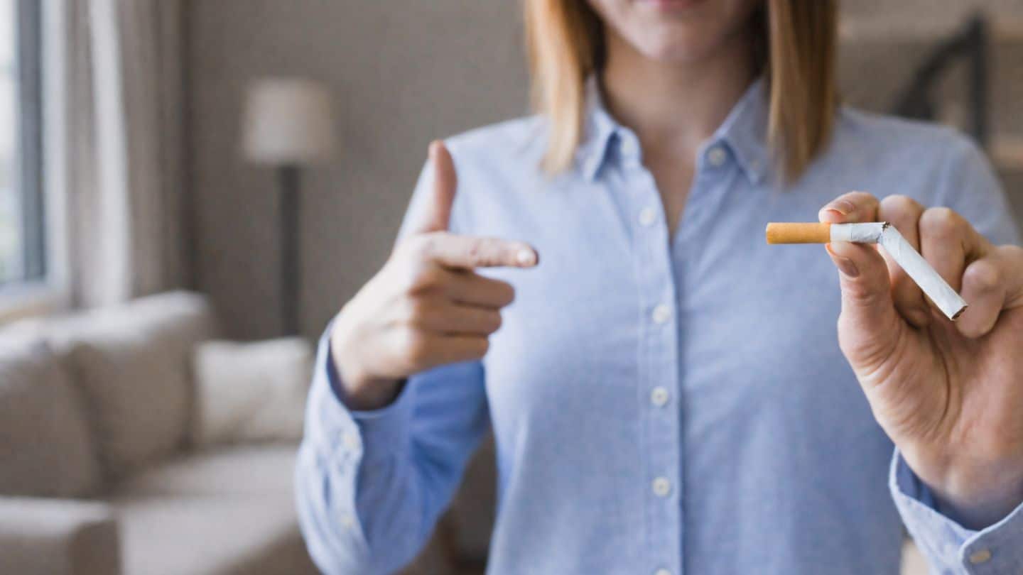Frau in hellblauem Hemd zeigt zerbrochene Zigarette und gibt Stopp-Geste mit der Hand, symbolisiert Rauchstopp