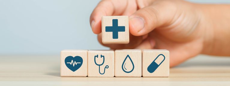 Hand platziert Holzwürfel mit Plus-Symbol auf Würfel mit Herz-, Stethoskop-, Wasser- und Pflaster-Icon
