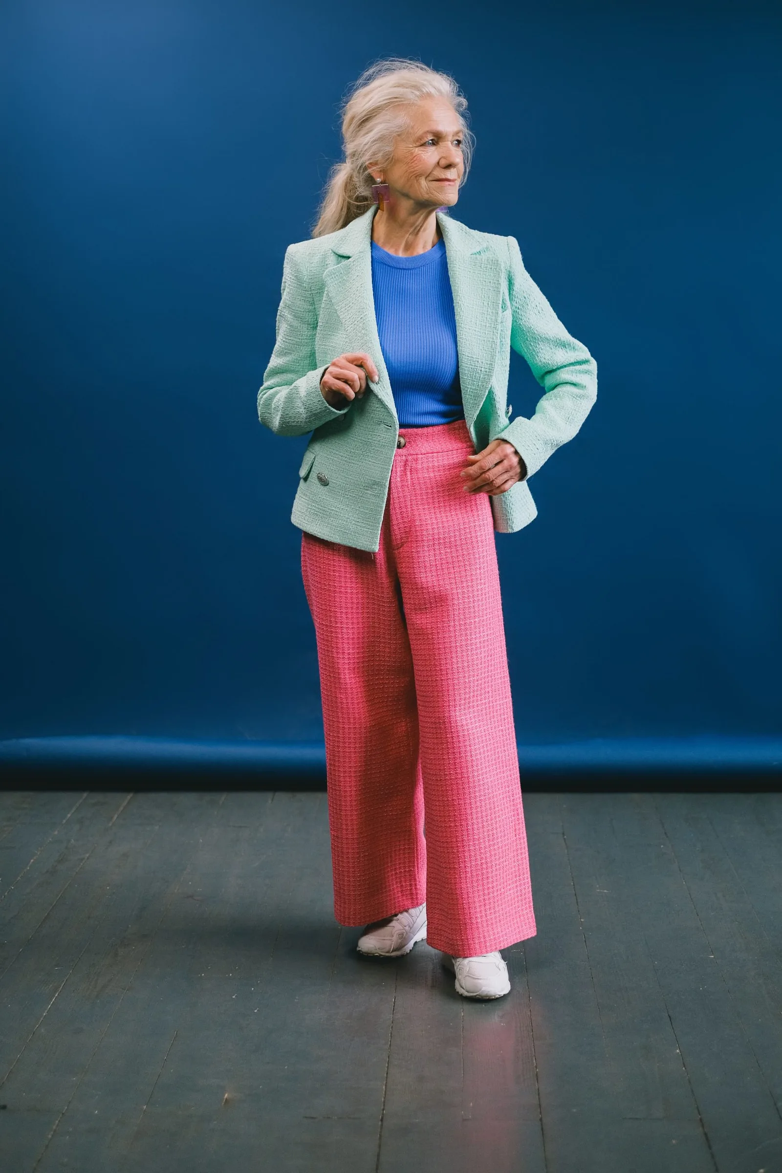 Ältere Frau mit blondem Haar im grünen Blazer, blauem Top und rosa Hose vor blauem Hintergrund