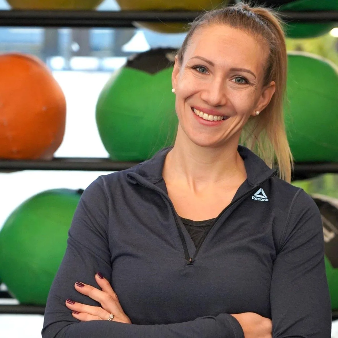 OTRANG – Trainer-Onlinekurs Rückbildung von Dr. Iris Floimayr-Dichtl 25 Porträt einer Frau in schwarzem Reebok-Shirt mit Fitnessgeräten im Hintergrund