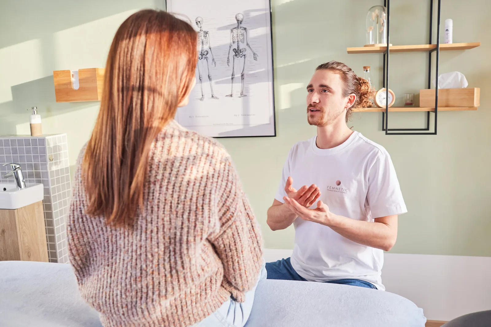 Mann in weißem T-Shirt mit Femnetic-Logo spricht mit Frau in beiger Strickjacke in modernem Behandlungsraum