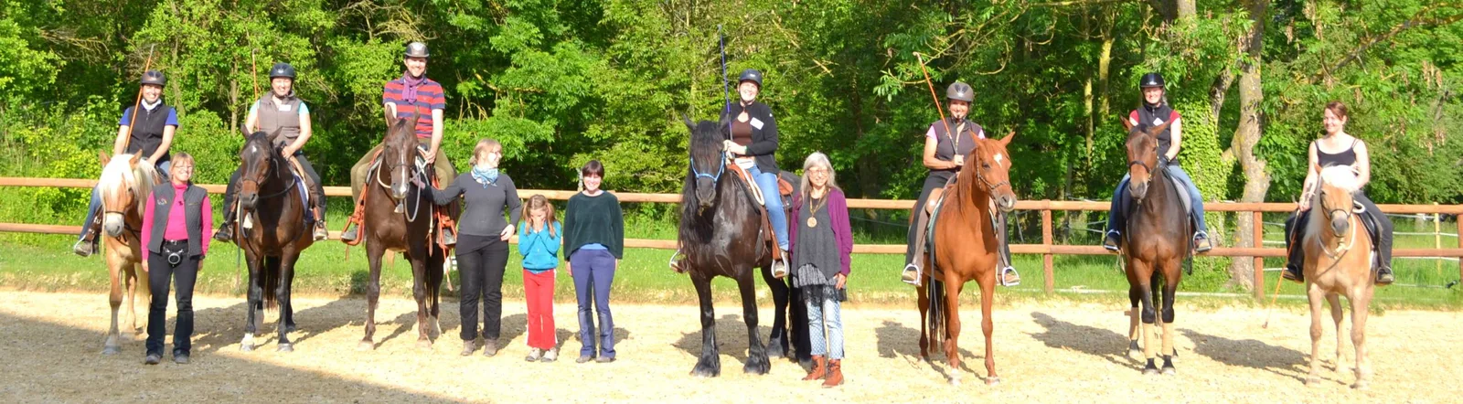 Gruppe von Reitern mit Pferden und Personen zu Fuß stehen in Reitplatz mit Zaun, grüne Wald-Kulisse