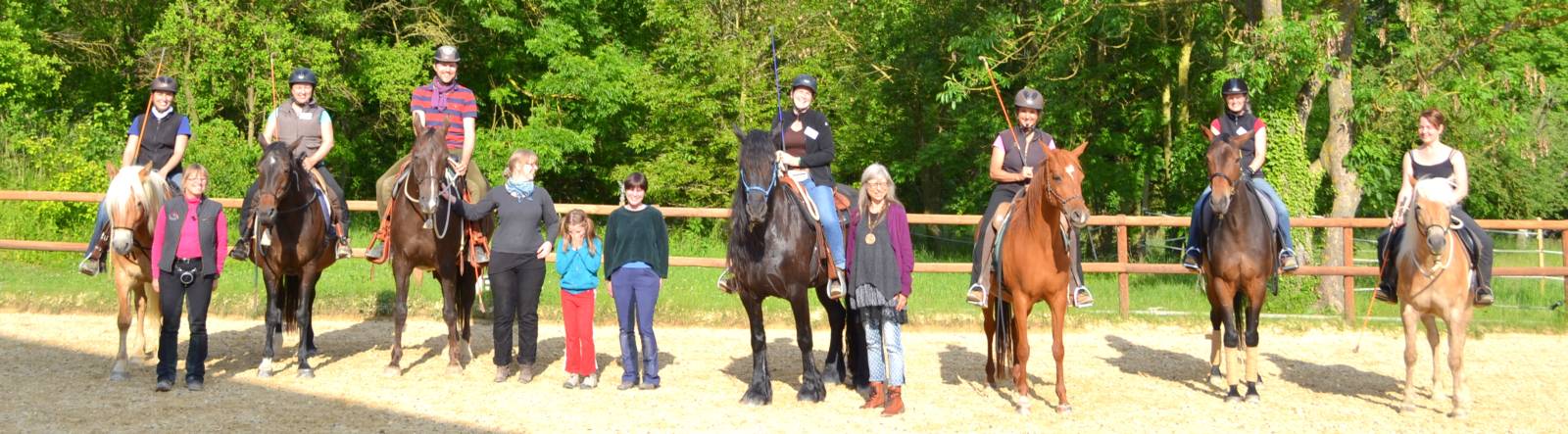 Gruppe von Reitern mit Pferden und Personen zu Fuß stehen in Reitplatz mit Zaun, grüne Wald-Kulisse