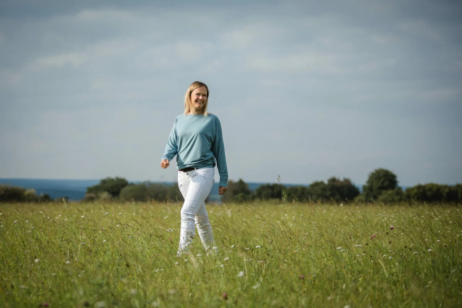 Junge Frau in hellblauem Pullover und weißer Hose läuft lächelnd durch blühendes Frühjahrsfeld mit Meerblick