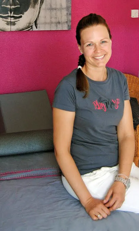 Physiotherapeutin Andrea in grauem PhyMayö Shirt sitzt lächelnd in pinker Praxis vor Matte