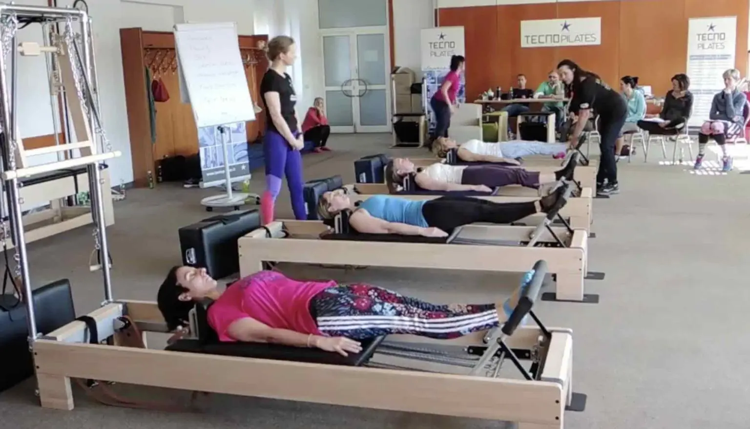 Pilates-Reformer-Kurs mit mehreren Teilnehmern in Liegeposition auf Geräten, Trainerin im Hintergrund