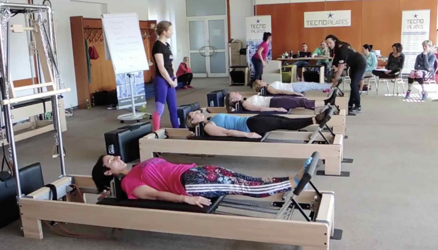 Pilates-Reformer-Kurs mit mehreren Teilnehmern in Liegeposition auf Geräten, Trainerin im Hintergrund