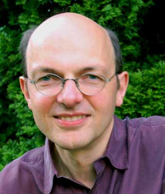 Porträt von Armin Risi, Mann mit Brille und violettem Hemd vor grünem Hintergrund